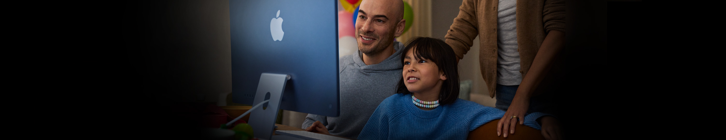Two people sitting together use an iMac while a third person stands behind them observing.